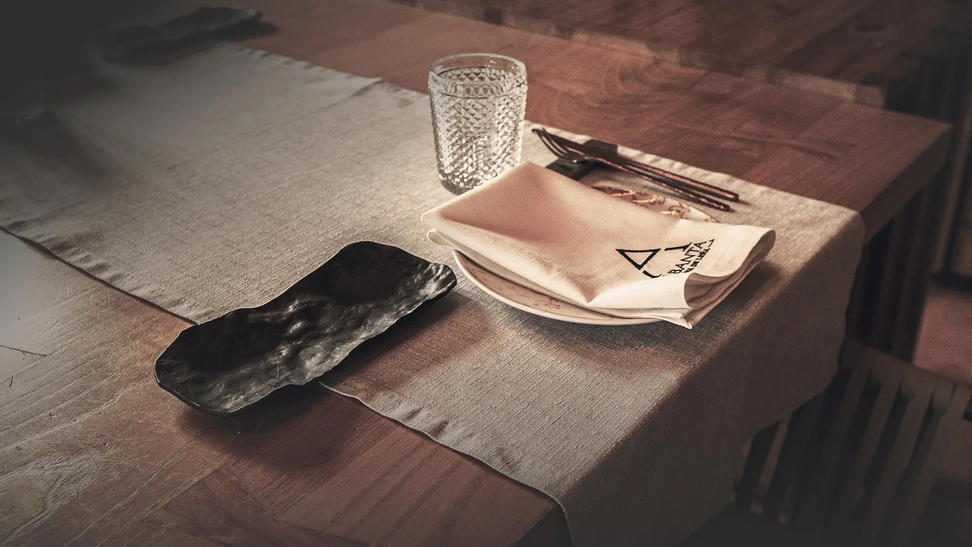 Mesa preparada en Albanta Cocina de Leña — servilleta bordada, vaso tallado y cubierto sobre pizarra oscura